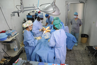 In a surgical suite, about five medical personnel surround a patient on a gurney.  A metal light is situated above the patient.  In the background, another medical person stands near the doorway. Medical equipment lines the back and left sides of the room.