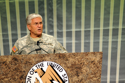 A man in a military uniform stands behind a lectern. The lectern has the words "Association of the United States Army" on it.  Vertical stripes adorn the wall behind him. The name tape on his uniform says "Casey."