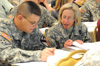 A man and a woman in military uniforms sit near each other at a table and appear to be studying.  Other soldiers can be seen in the background.
