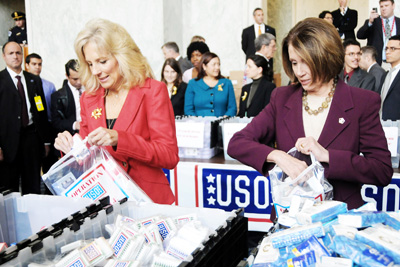 Two women pack goods into plastic bags.  Behind them is a sign that says "USO."  Dozens of other people are also in the room standing nearby.