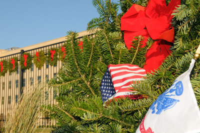 A wreath made of greenery has a red ribbon on it, an American flag and a U.S. Army flag. In the background is the Pentagon, seen through a black fence, which also has wreaths on it.