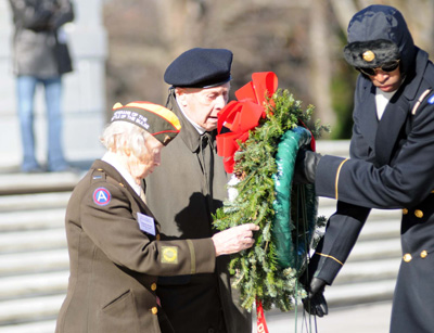 An elderly woman in a military uniform, wearing a hat that indicates her status as a veteran, along with an elderly man wearing a beret, place a wreath.  A Soldier helps them move the wreath.
