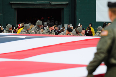Individuals in uniform gather around a huge flag, stretching it out over a football field.