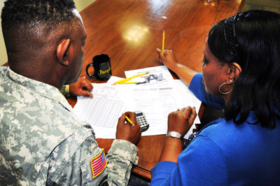 A man in a military uniform and a woman in a blue dress sit together at a table and review what appears to be financial information.  On the table are papers, a calculator and some writing implements.