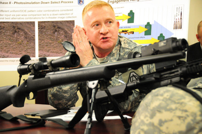 A man in a military uniform sits at a table.  On the table are several military rifles. In the background on the wall are charts and diagrams.