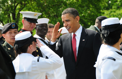 Outdoors, a woman in a military uniform salutes a man in a suit.  Other military personnel are gathered around nearby.
