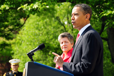 Outdoors, a man in a suit stands behind a lectern.  A woman stands nearby.  In the background, a man is in a military uniform.
