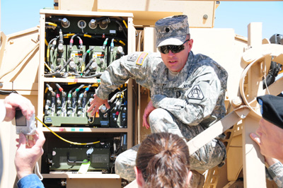 A man in a military uniform has opened a hatch on a combat vehicle and is displaying cabling and electronics inside.  A few people are gathered around and are watching. One takes a photo.