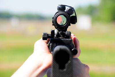 Two hands hold a rifle, and both a red dot and the iron sights can be seen though the rifle's scope.