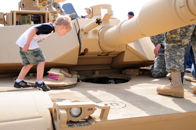 A small boy stands on top of a tank and peers down into an open hatch.