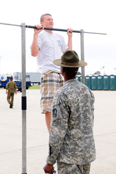 A young man does pull-ups on a bar.  A soldier stands nearby and watches.  In the background are portable toilets.