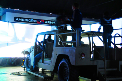 A mock up of a combat vehicle sits indoors in a theater-like environment.  A large projection screen is at the front of the room displaying a simulated combat situation the vehicle might be involved in. A sign reads "America's Army."