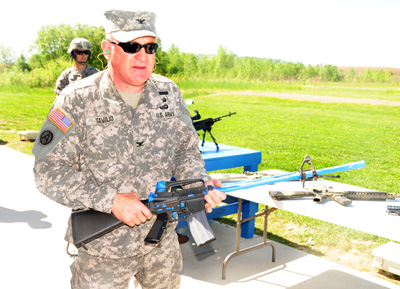 Outdoors at the edge of a field, a man in a military uniform holds a rifle that has various parts covered in blue tape.  In the rear is another solider and also a gun sitting on a table.  