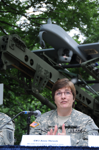 A woman in a military uniform sits outdoors at a table and speaks into a microphone. Behind her is a launch system for an unmanned aerial system.  In front of her a name tag says "CW3 Anne Thrush."
