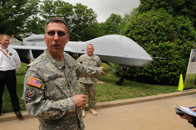 Outdoors in an area with trees and bushes, a man in a military uniform points behind him at a static display of an unmanned aerial vehicle. Another soldier stands next to the vehicle, as does a civilian.