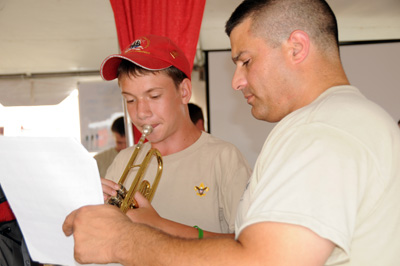 A boy in a red hat plays a musical instrument.  A man in a beige T-shit holds music up for the boy.