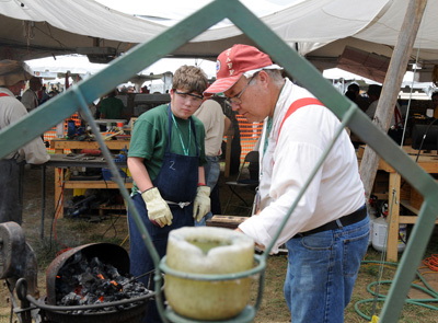 Outdoors, an older man in a red hat uses a poker to manipulate coals in a furnace.  A boy wearing an apron stands nearby and watches.  In the rear is a tent with other people operating tools and equipment.