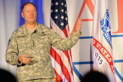 A man in a military uniform stands on a stage. Behind him are the American flag and the U.S. Army flag.
