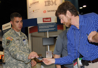 A man in a military uniform hands something to a man in a blue striped shirt.  In the background are displays indicative of a conference.  One reads "IBM."