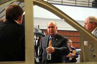 As seen through the door frame of a yet unfinished combat vehicle, a man in a suit talks to another man in a suit.  Others men stand nearby.  