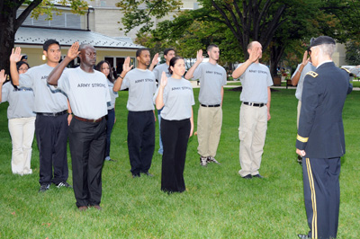 Outdoors on a lawn, about a dozen individuals in T-shirts that say "Army Strong" stand in a formation and raise their right hand.  A military officer stands in front of them and administers an oath.
