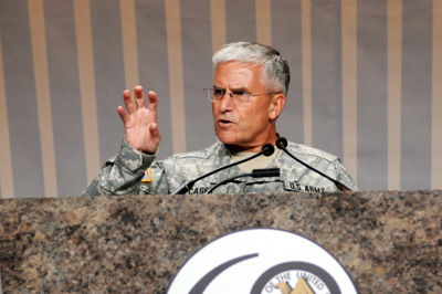 A man in a military uniform stands behind a huge lectern.  His right hand is in the air making a claw shape.  