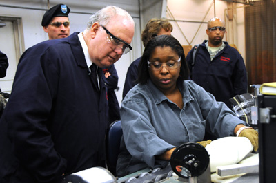 A woman sits at a bench and assembles a bomb.  A man in a suit stands near her and watches.  Other people are in the background watching them interact.