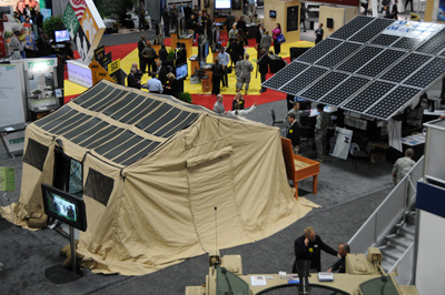 A tent set up indoors has solar panels on top. Nearby are other solar panels and other displays, and crowds of people.
