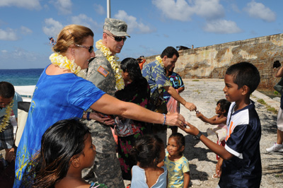 Outdoors on a beach, a woman places something into the hand of a boy.  Nearby a man in a military uniform watches. He is surrounded by small children.  In the background, a man in a floral shirt also hands something to a child.