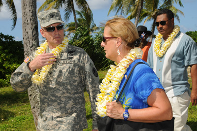 A man in a military uniform and wearing a lei talks with a woman also wearing a lei.  The two are walking outside.  Palm trees are in the background. Another man follows behind. 