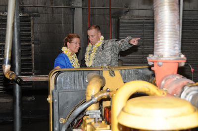 A woman wearing a lei and a man in a military uniform, also wearing a lei, are inside an industrial shed near what appears to be a generator.  The man in the uniform points at the generator.