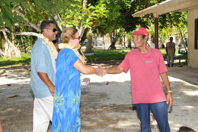 In an outdoors tropical setting, a woman in a blue dress and wearing a lei shakes hands with a man in a red shirt and red hat. Another man stands next to the woman.  