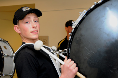 A young man in a black shirt carries a large drum. He holds drum sticks in his hands.  Behind him another drum can be seen.