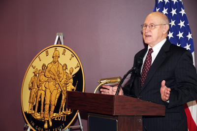 A man in a suit stands behind a lectern.  To his right is a large mock-up of a gold coin. It says "Liberty - 2011 - In God We Trust." Behind the man is an American flag.