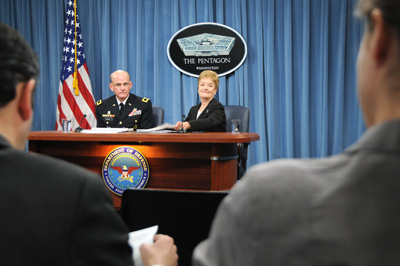 A man in a military uniform and a woman in a black suit sit behind a wooden table with a Department of Defense seal on the front. Behind them is an American flag, blue drapes, and sign that has an image of the Pentagon on it which reads "The Pentagon - Washington."  Other people sit towards the front of the room and look at them.