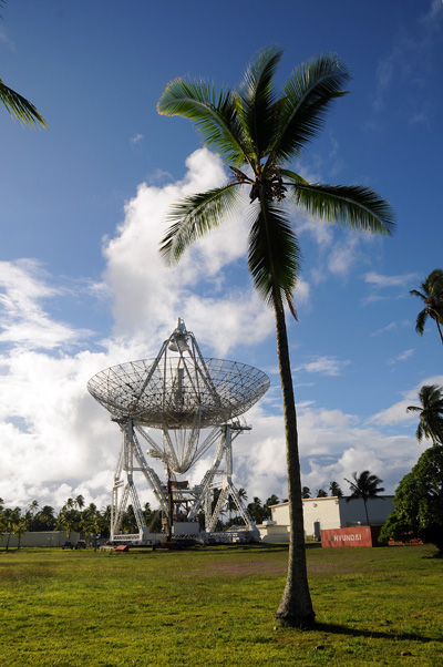 Near a short building, a radar dish points towards a blue sky with puffy white clouds.  Palm trees are seen all around. A cargo container reads "Hyundai."