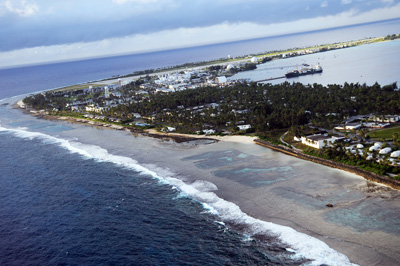 From a high altitude, water from the ocean is seen washing up on an island that has buildings and radar domes.