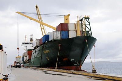 A cargo ship is docked at a port.  It holds multi-colored containers.  Several cranes are mounted to the surface of the ship.  One cargo container is being lowered to the dock.