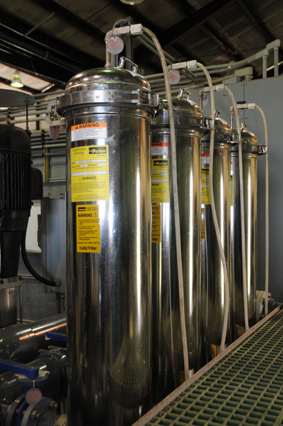 A room has four stainless steel canisters with tubing coming out from the top. On the right, a grated floor.  In the background is other equipment.