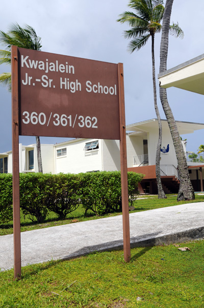 A brown sign reads "Kwajalein Jr.-Sr. High School."  Palms and buildings are in the background.