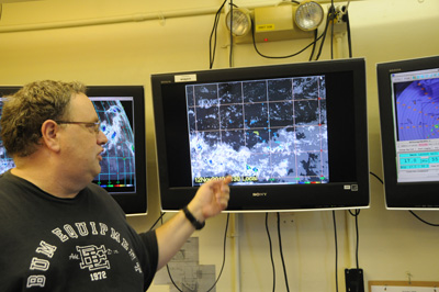 A man points to one of three television screens on a wall.  They appear to display weather satellite information. 