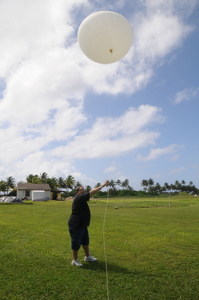 In the middle a field, a man launches a weather balloon.  In the background is a small building and a tree line of palms.  
