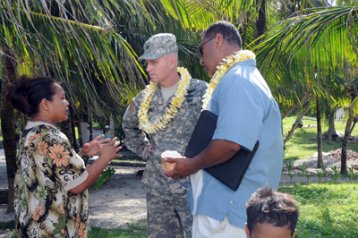 A man in a military uniform weirs a lei around his neck.  He talks with a woman in a floral dress.  Palm trees surround them. A civilian man in a blue shirt also wears a lei.