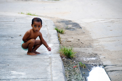 A small boy wearing only a diaper squats on a sidewalk and picks up a stone.