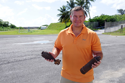 A man in an orange shirt holds what appear to be two small bombs.  He stands in a parking lot.