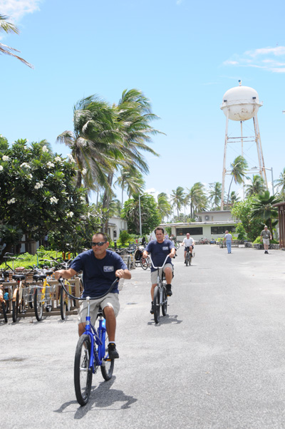 In a tropical setting, multiple individuals ride bicycles on a paved road.  A water tower stands in the background amidst palm trees and other buildings.