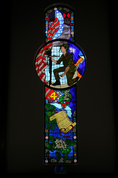 A stained glass window depicts a soldier down on one knee, a U.S. flag, and other tropical elements.