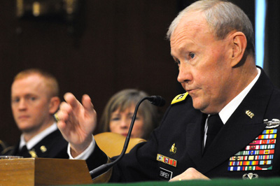 A man in a military uniform with many ribbons sits and speaks into a microphone.  Two other people are behind him and to his right.