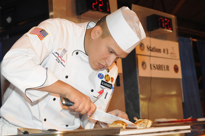 A young man in a chef's outfit uses a large knife to cut food.  Behind him are signs on a refrigerator that say "Station #3" and "USAREUR."
