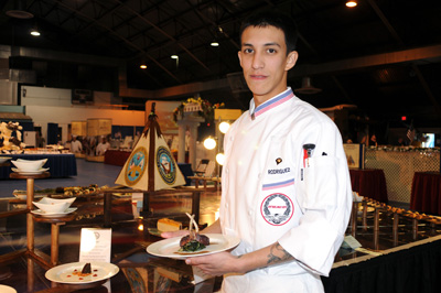 A young man in a chef's outfit holds a plate of food.